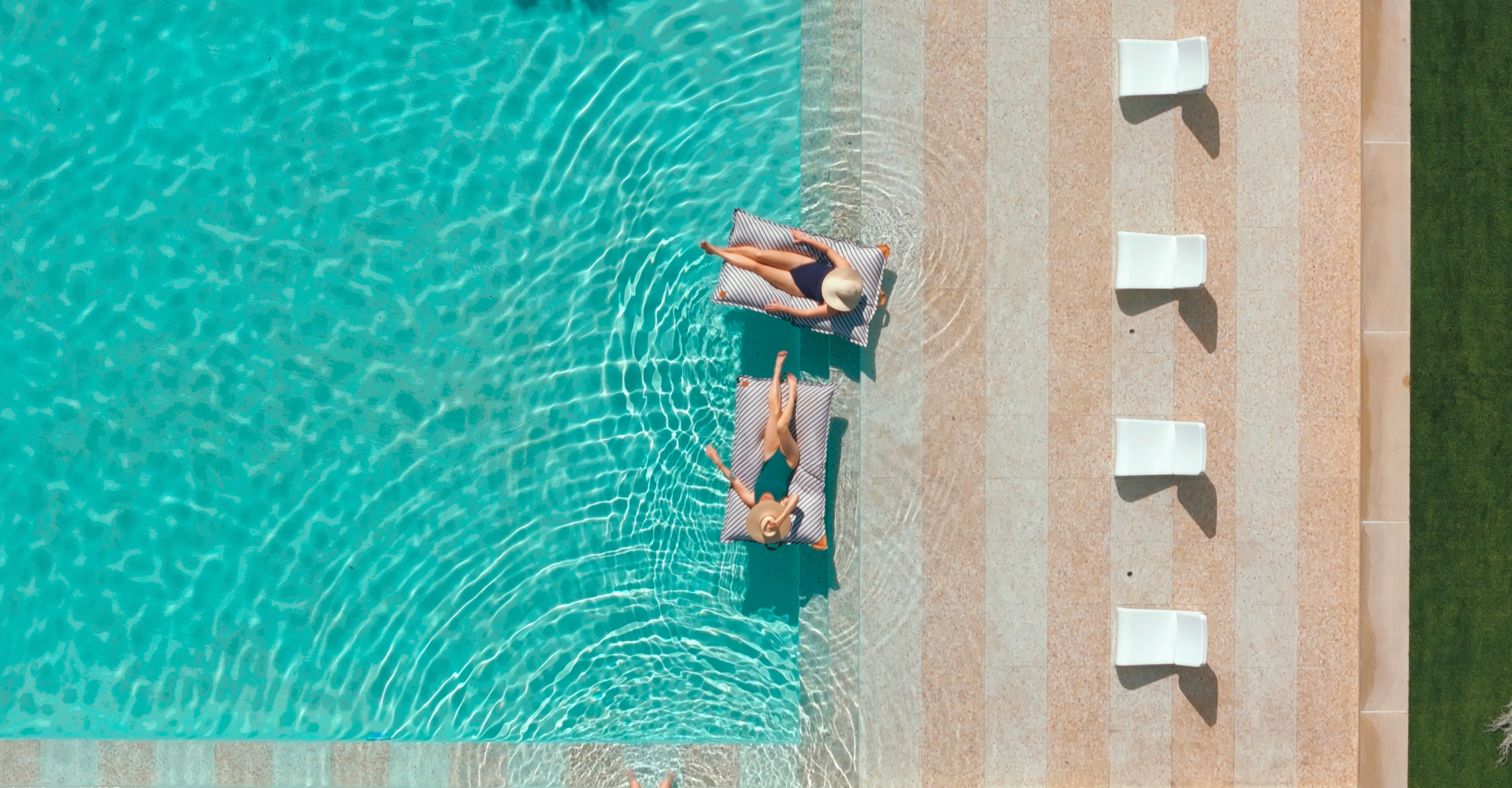 Two people lying on lounge chairs by a poolside with clear blue water.