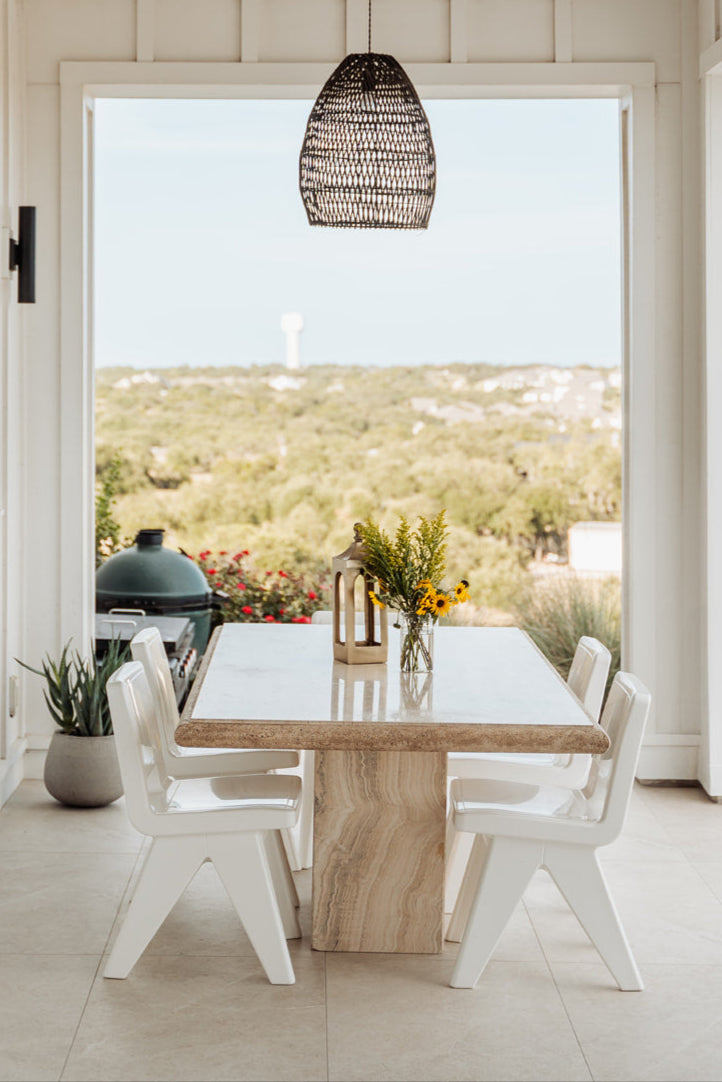 Dining area with a wooden table and chairs, large window view of greenery, and hanging wicker lamp.